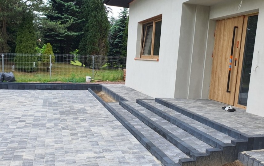 Ułożona szara kostka brukowa na podjeździe i schodach przed nowoczesnym domem z drewnianymi drzwiami i oknem, widoczny fragment ogrodu z siatką ogrodzeniową.