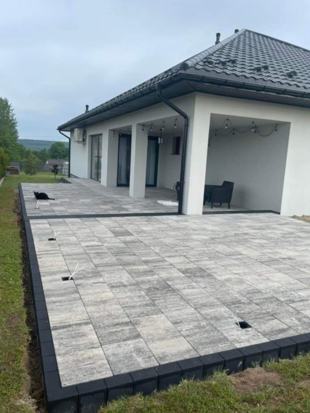 Wyłożony kostką brukową taras z czarną obwódką, przy nowoczesnym domu z oświetleniem, na którym siedzi czarny kot.