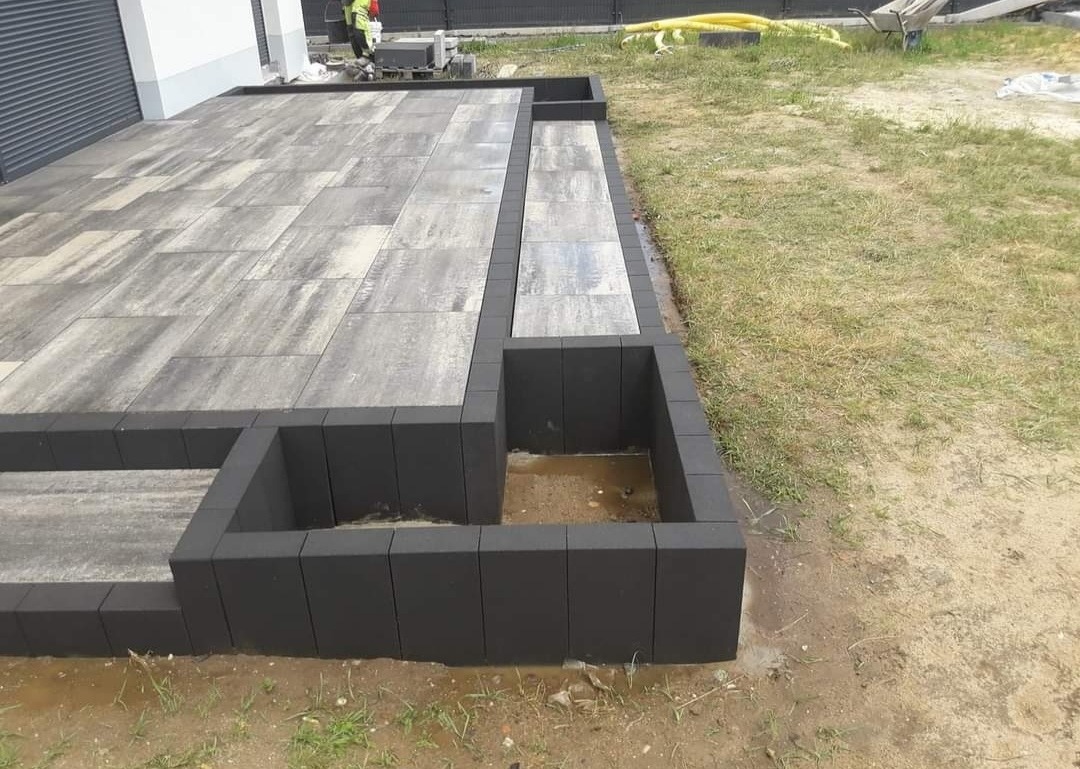 Wykończony taras z szarej kostki brukowej z elementami podwyższonymi obłożonymi czarną kostką, trawnik w tle.