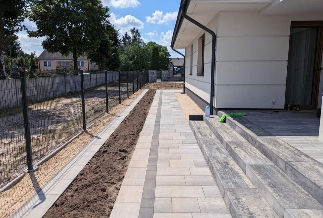 Nowoczesna posesja w Kielcach: świeżo ułożona kostka brukowa tworząca ścieżkę i schody wejściowe, obok przygotowany teren pod nasadzenia, metalowe ogrodzenie panelowe w tle.