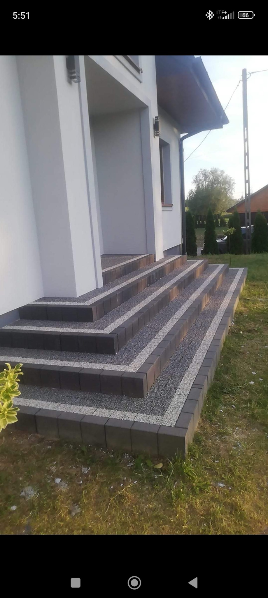 Schody wejściowe do domu wyłożone ciemną kostką brukową z dekoracyjnym wypełnieniem z jasnego grysu, otoczone zielonym trawnikiem.