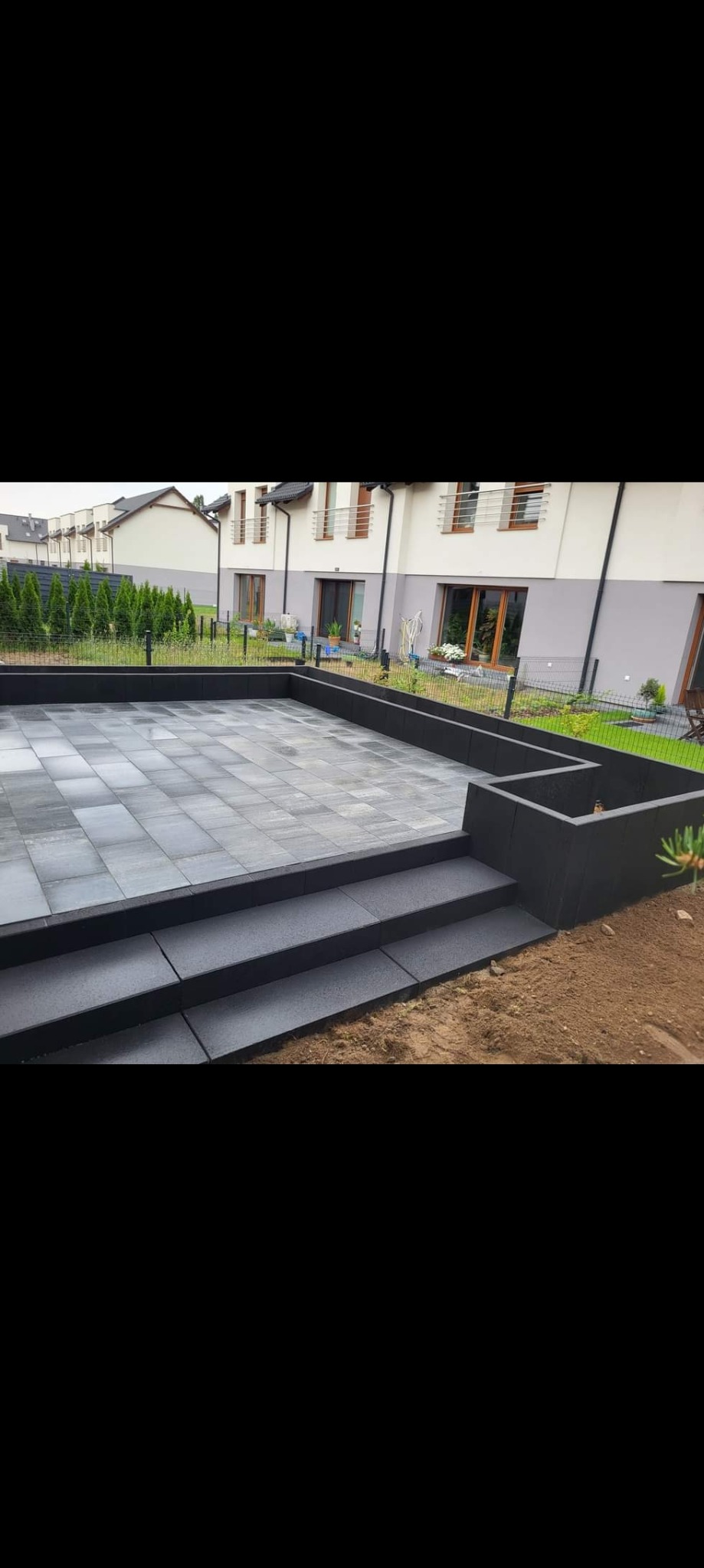 Nowoczesny taras z szarej kostki brukowej, otoczony czarną obudową, z trzema schodami wejściowymi, na tle domu z jasną elewacją i ogrodzeniem.
