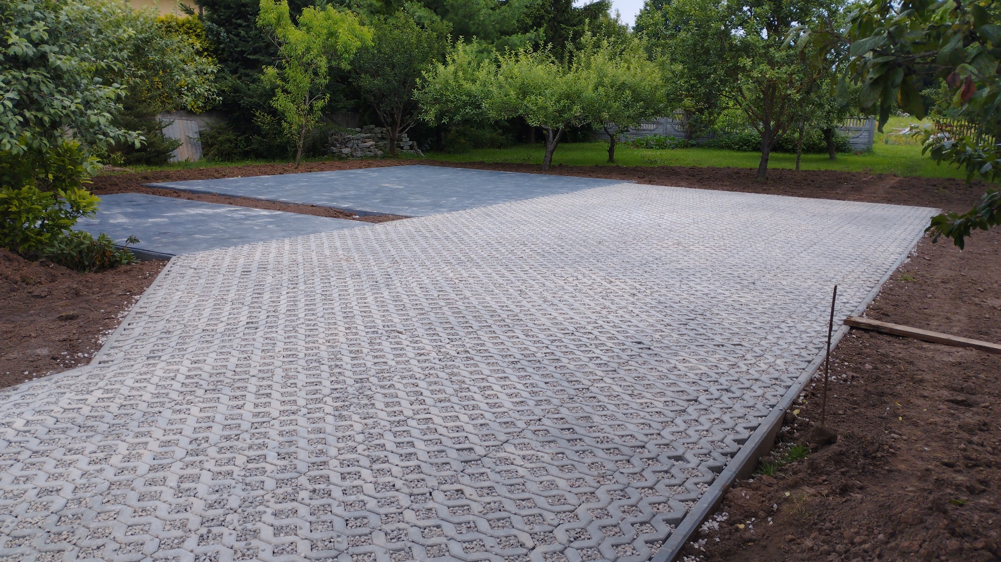 Ułożona szara ażurowa nawierzchnia z wypełnieniem z drobnych kamyczków, obrzeża z metalu, w tle przygotowane podłoże pod dalszą część nawierzchni, teren zielony.