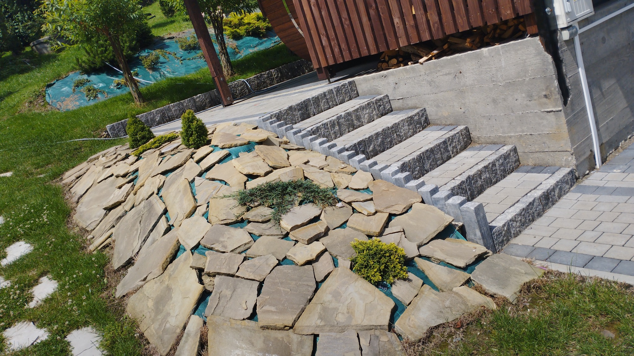 Ułożone nieregularne płyty kamienne na skarpie z roślinami, obok schody z kostki brukowej prowadzące do drewnianej konstrukcji.