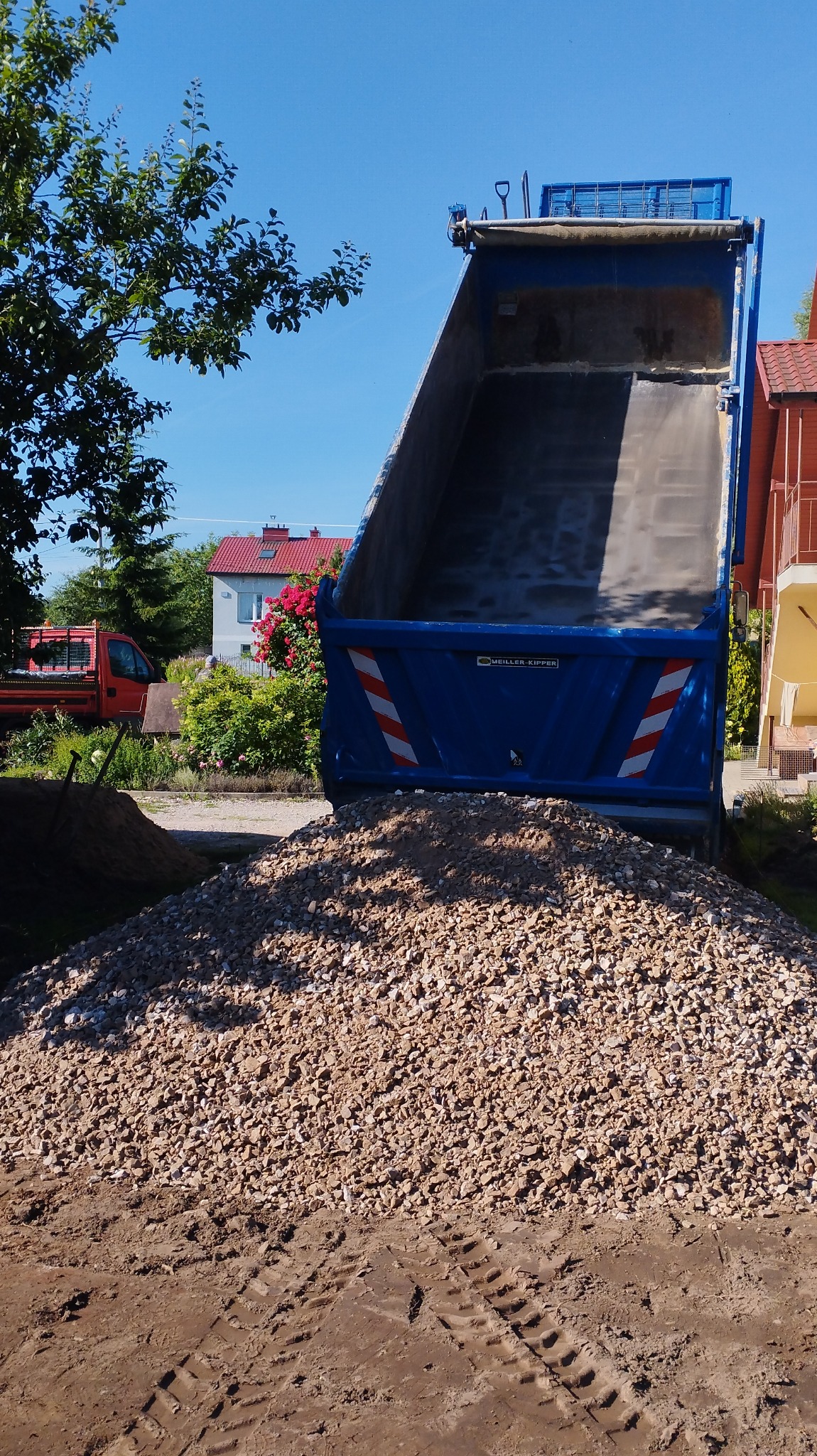 Wyładowany z wywrotki granulat kamienny na ziemi z widocznymi śladami opon, w tle zabudowa jednorodzinna i zieleń.