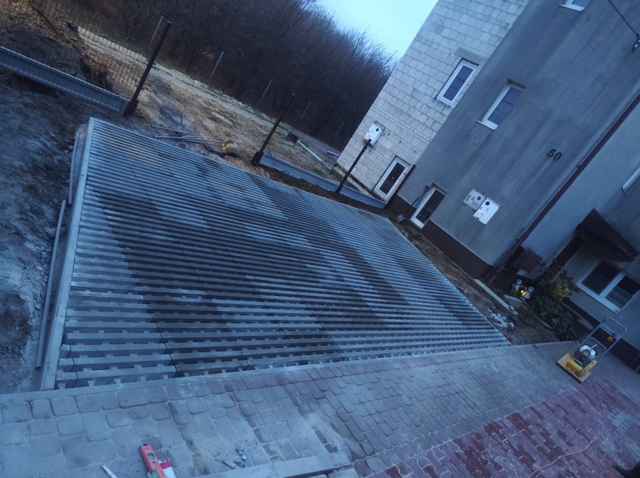 Ułożona, szara kostka brukowa w pasy, z widocznym fragmentem budynku z numerem 50 i ubijarką. Częściowo ułożona kostka czerwona na schodach.