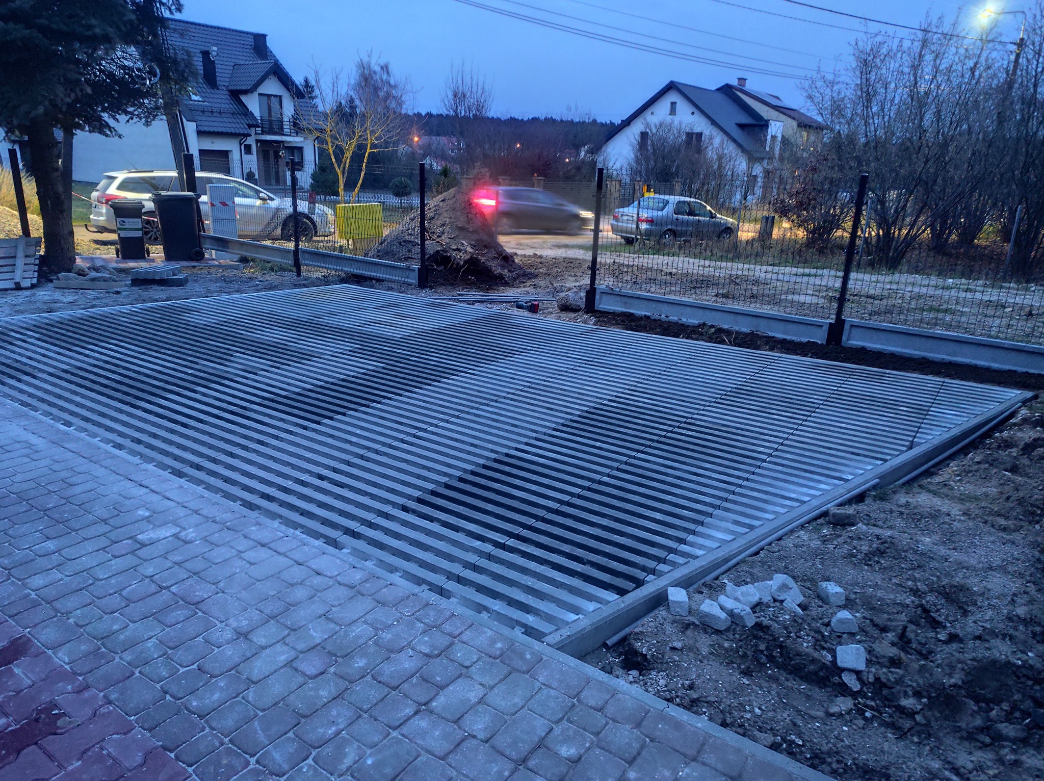 Szara, ażurowa konstrukcja z betonu na posesji, prawdopodobnie podjazd lub parking. W tle domy i samochody, widoczne elementy ogrodzenia. Zdjęcie wykonane o zmierzchu.