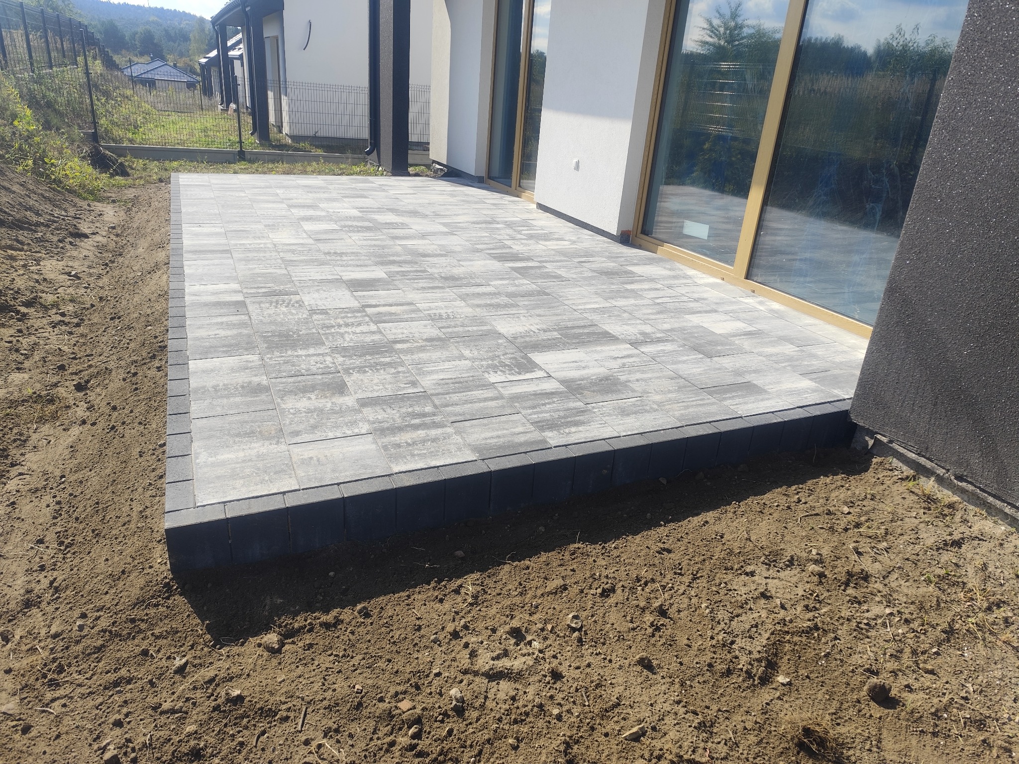 Nowoczesny taras z szarej kostki brukowej, obrzeża z ciemnej kostki, widok na dom z dużymi oknami, surowy grunt wokół tarasu.