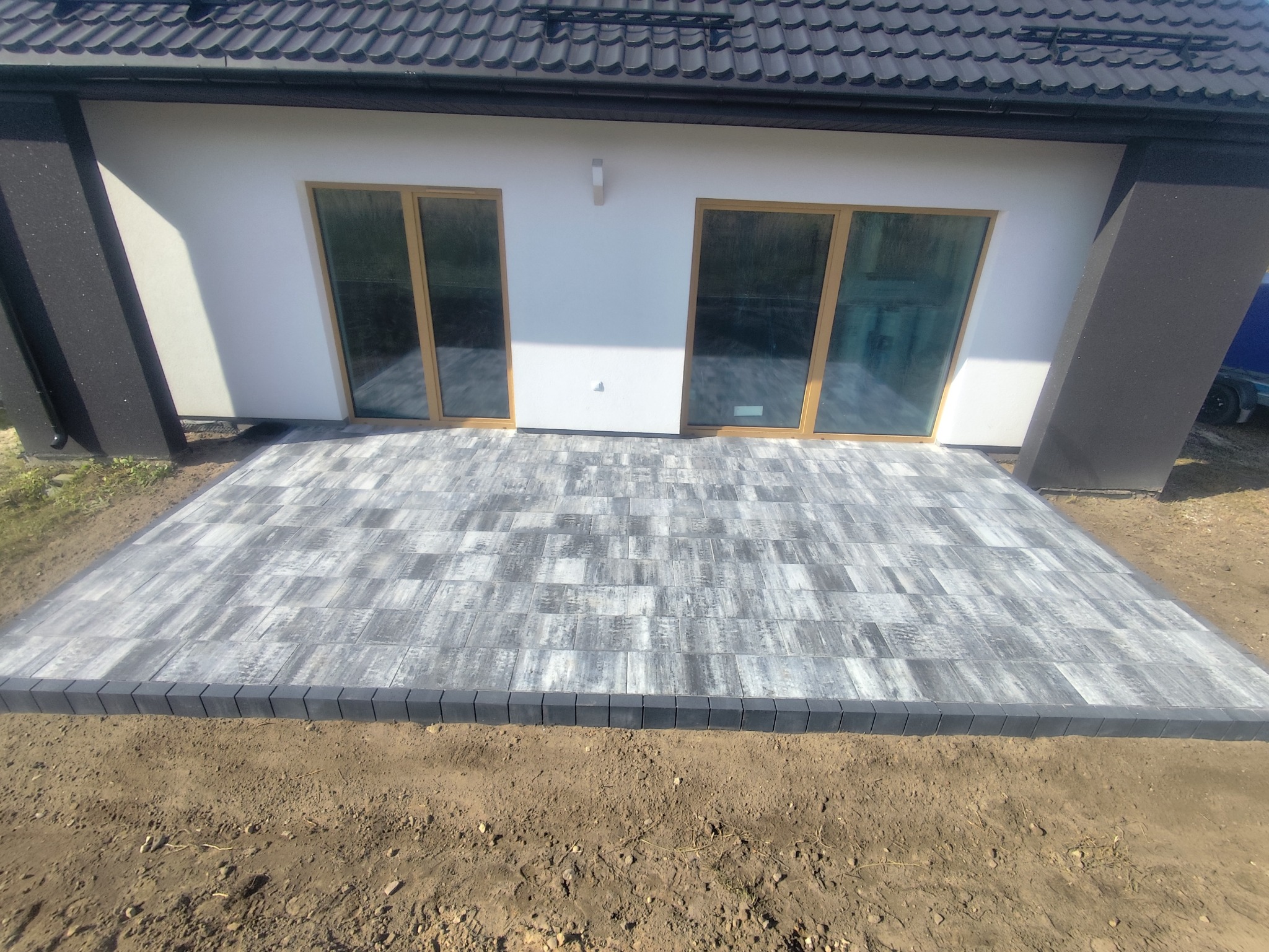 Nowoczesny taras z szarej kostki brukowej ułożonej wzorem prostokątnym, obrzeża z ciemnej kostki. Tło stanowi biała elewacja budynku z dużymi oknami.