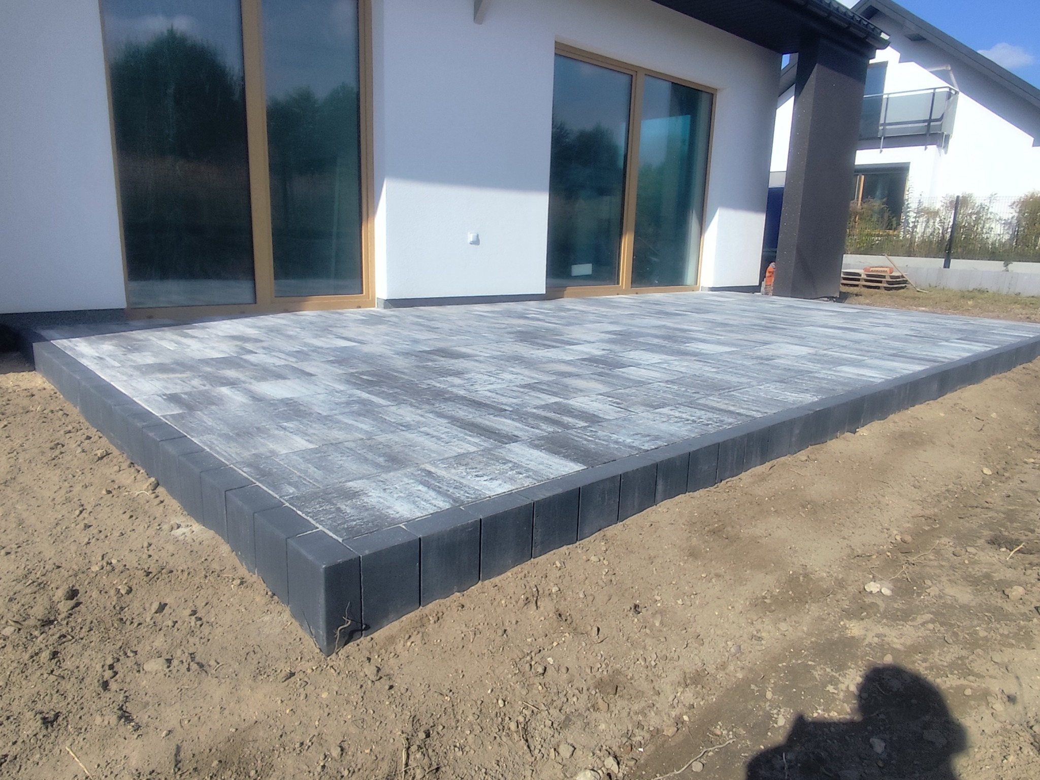 Nowoczesny taras z szarej kostki brukowej, obrzeża z ciemnych kostek. Widok na biały budynek z dużymi oknami. Minimalistyczny design, surowe wykończenie.