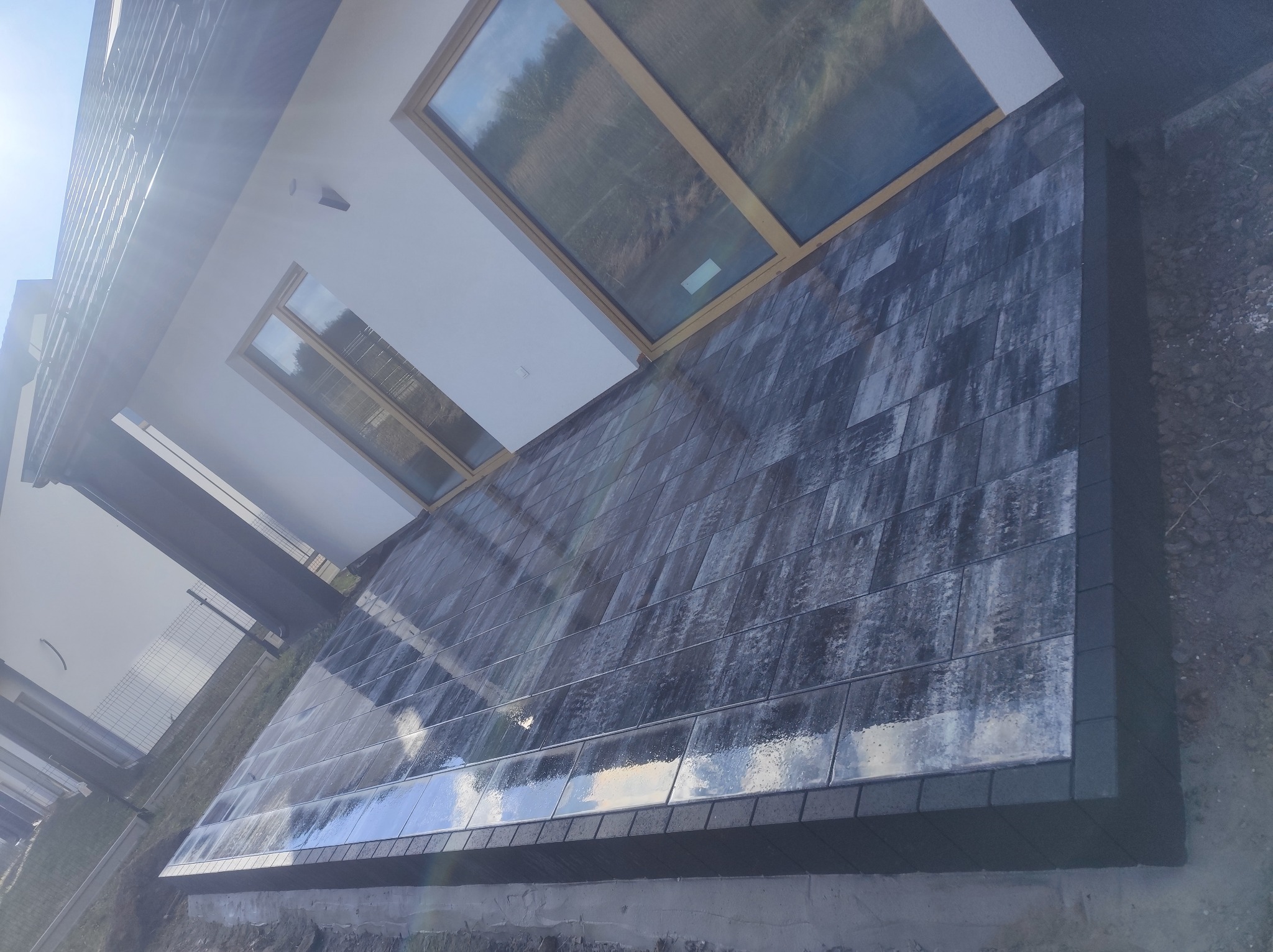 Nowoczesny taras z ciemnej kostki brukowej, widok z dołu, z refleksami światła na powierzchni. Minimalistyczny design, okna dachowe w tle.