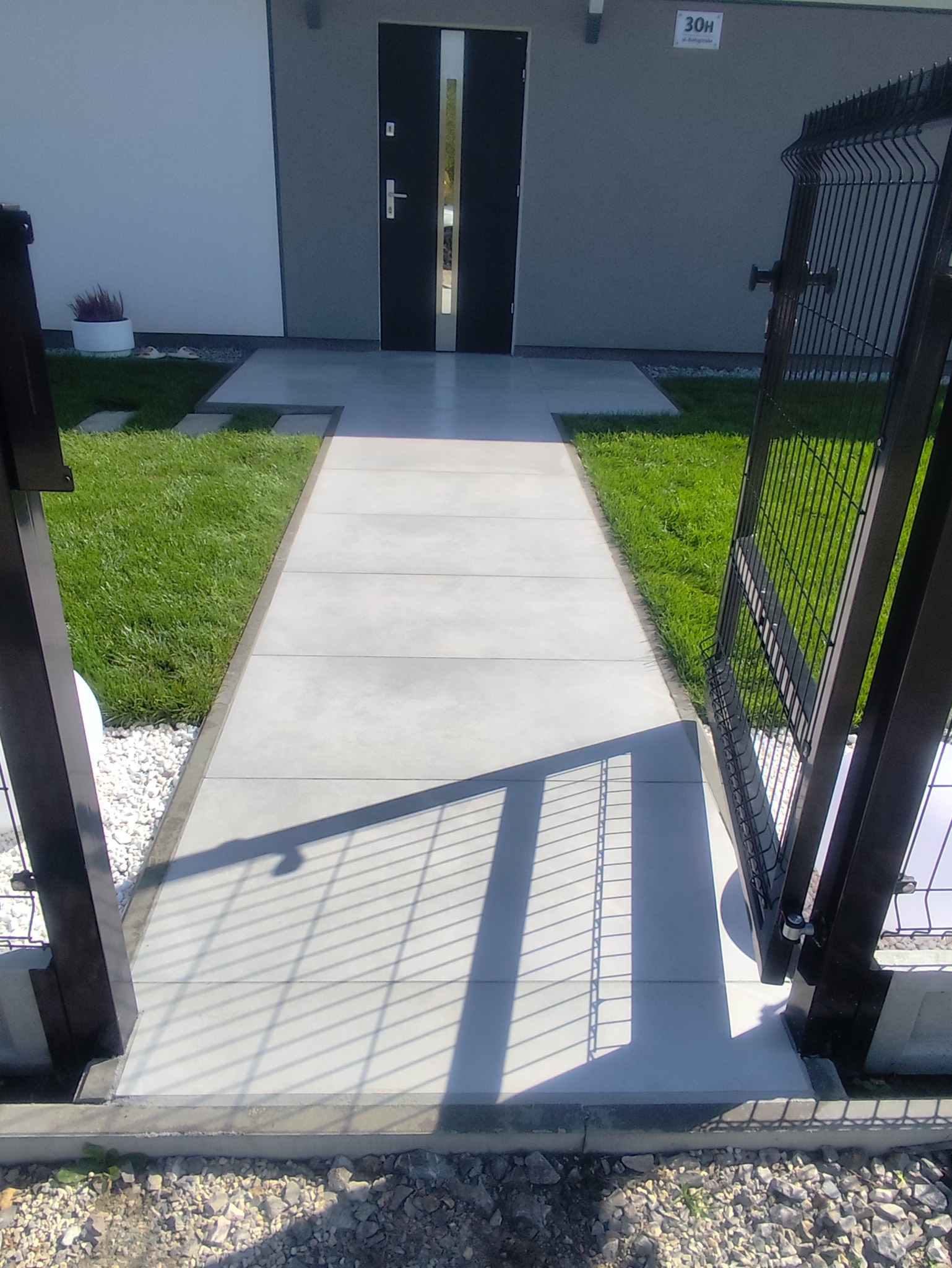 Nowoczesny chodnik z szarych płyt prowadzący do drzwi wejściowych, obok trawnik i otwarte metalowe ogrodzenie. Widok z perspektywy.
