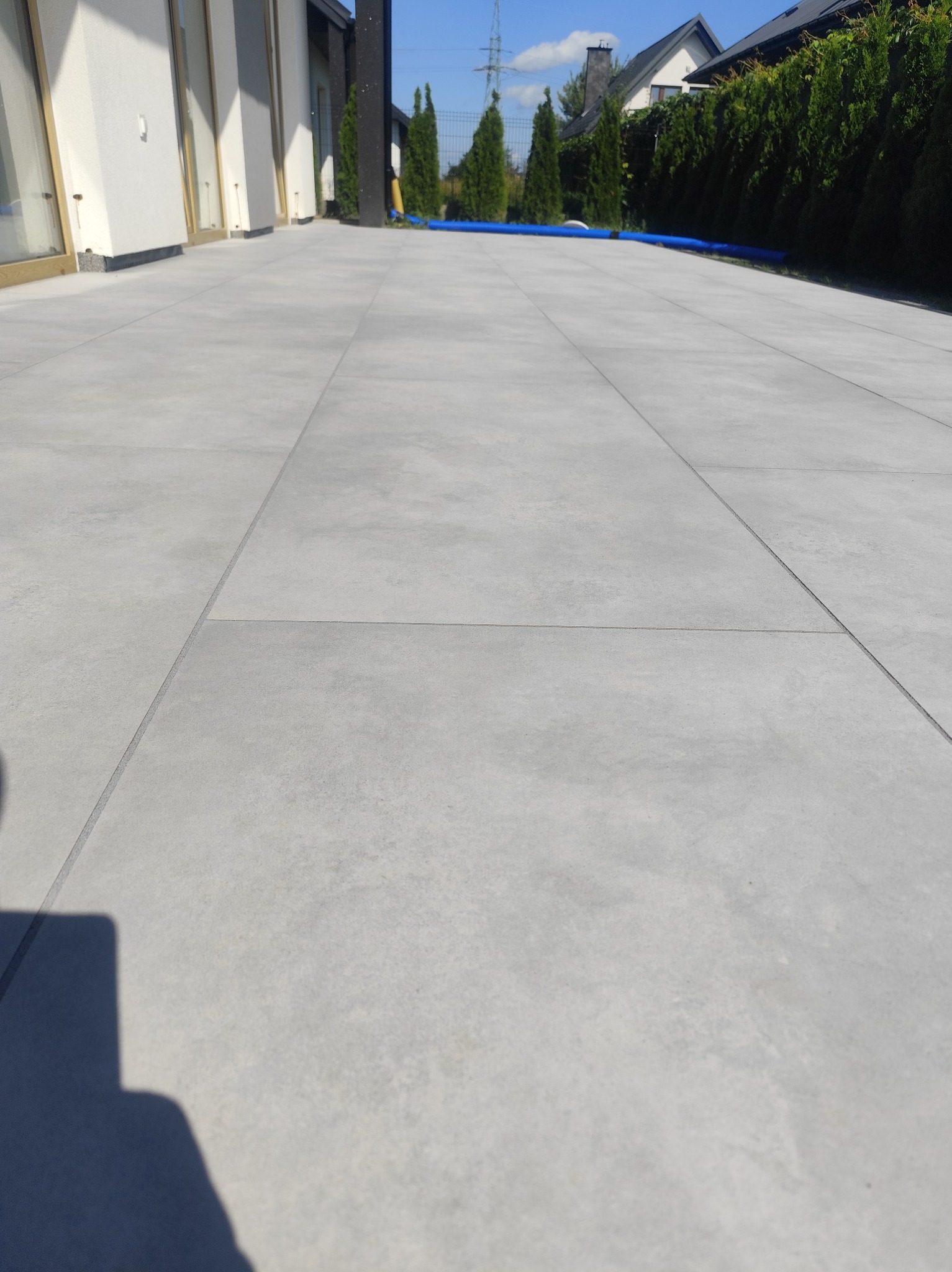 Nowoczesny taras wyłożony dużymi, szarymi płytami. Widoczne geometryczne linie fug, budynek w tle i fragment niebieskiego węża ogrodowego.
