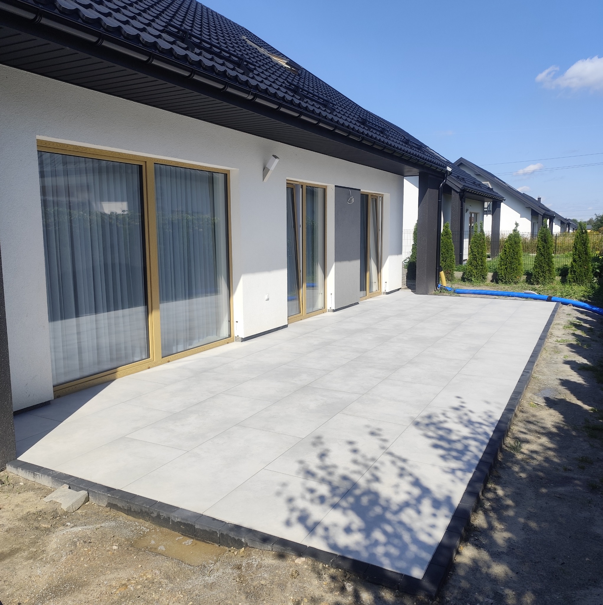 Jasnoszary taras z dużych płyt, obrzeża z ciemnej kostki, przylegający do białego domu z czarnym dachem. Błękitne niebo w tle. Widoczne złącza płyt.