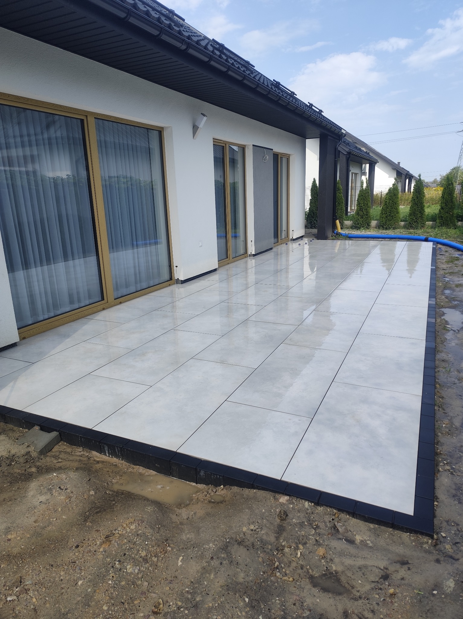 Jasnoszara, prostokątna kostka brukowa ułożona na tarasie przylegającym do białego domu z dużymi oknami. Czarna obwódka kostki. Widoczne ślady po deszczu.