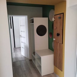 Budomixs - Przedpokój z białymi meblami: szafką z półkami i okrągłym otworem, drewnianym panelem ściennym z wieszakami, podłogą z paneli imitujących drewno, ściany w odcieniach zieleni i żółci.