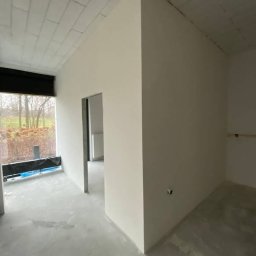 Budomixs - Wnętrze w trakcie prac wykończeniowych: gładkie, jasne ściany po szpachlowaniu i malowaniu, surowy beton na podłodze, widok na zewnątrz przez okno z roletą.