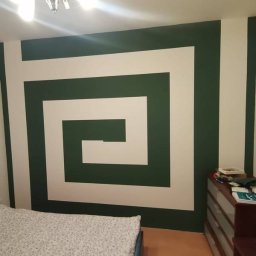 Budomixs - Ściana w sypialni z geometrycznym wzorem w kolorach zieleni i bieli, nad łóżkiem z wzorzystą pościelą, oświetlona przez lampę sufitową z kilkoma żarówkami.