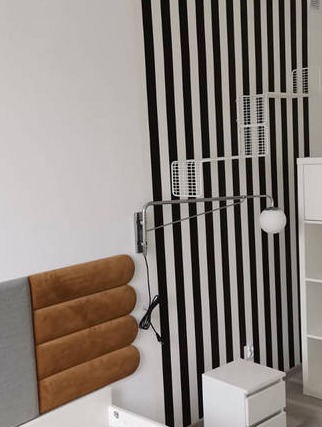 Pokój z tapetą w czarno-białe pasy, regulowaną lampą ścienną z kulistym kloszem, panelem tapicerowanym w odcieniach brązu i szarości oraz białymi meblami