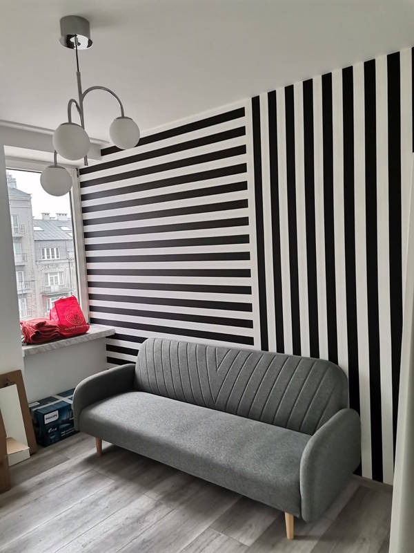 Pokój z szarą sofą na tle ściany w czarno-białe pasy, widoczna lampa sufitowa z trzema kulistymi kloszami i okno z widokiem na budynki.