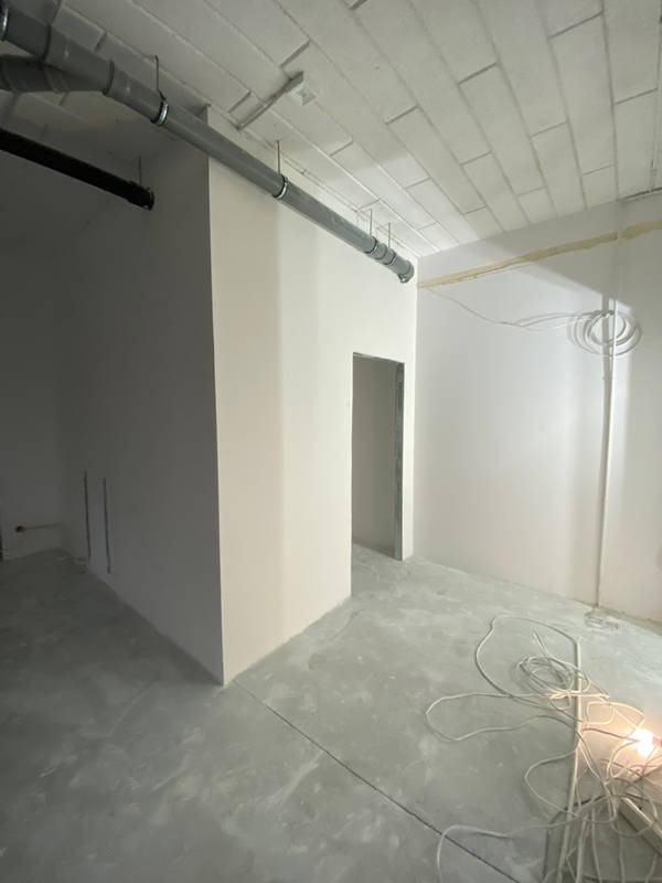 Wnętrze w stanie deweloperskim z surowym betonem na podłodze, świeżo pomalowanymi ścianami na biało, widocznymi instalacjami pod sufitem i kablem elektrycznym leżącym na podłodze.