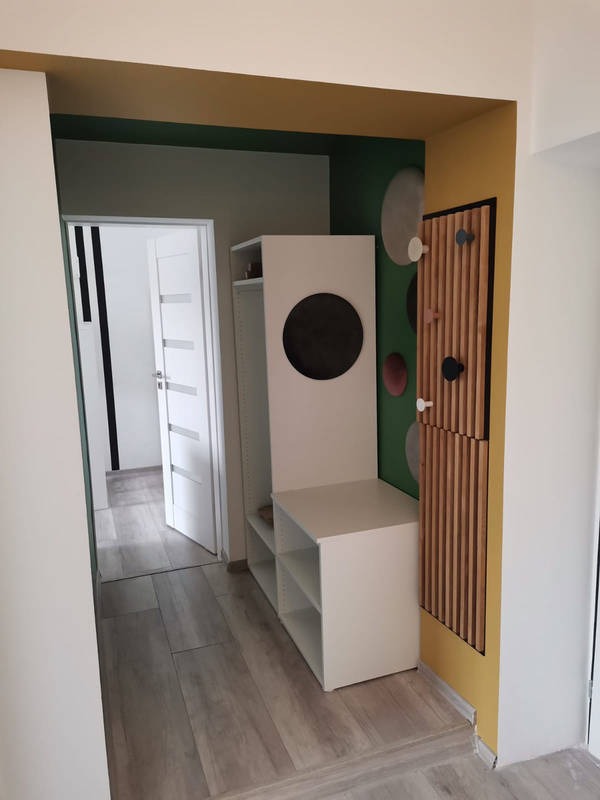 Przedpokój z białymi meblami: szafką z półkami i okrągłym otworem, drewnianym panelem ściennym z wieszakami, podłogą z paneli imitujących drewno, ściany w odcieniach zieleni i żółci.