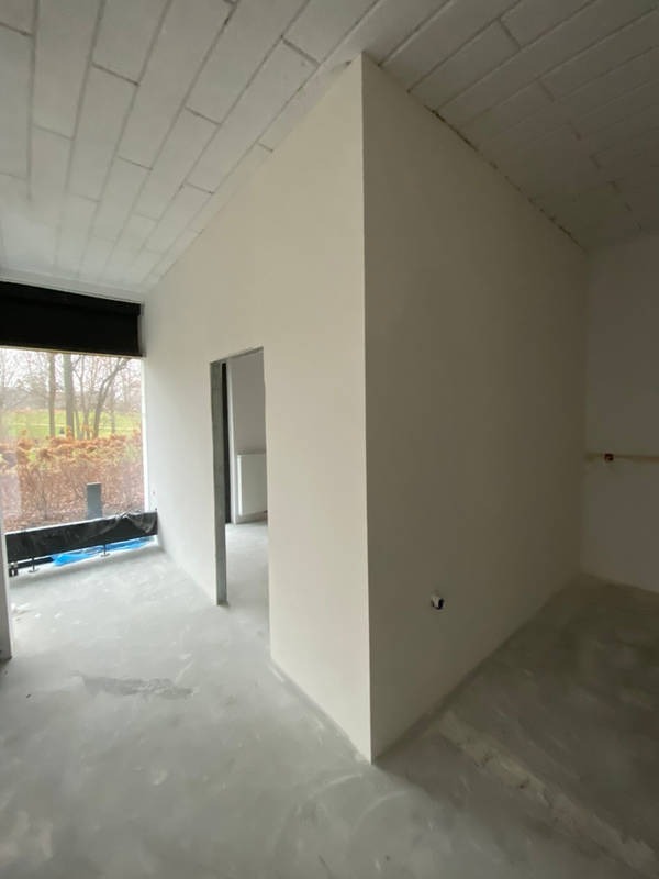 Wnętrze w trakcie prac wykończeniowych: gładkie, jasne ściany po szpachlowaniu i malowaniu, surowy beton na podłodze, widok na zewnątrz przez okno z roletą.