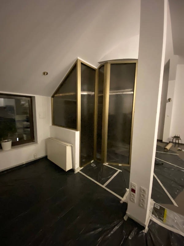 Pokój z przygotowaną podłogą zabezpieczoną czarną folią i taśmą malarską, widoczna przeszklona konstrukcja z złotymi ramami, grzejnik pod oknem i białe ściany w trakcie prac.