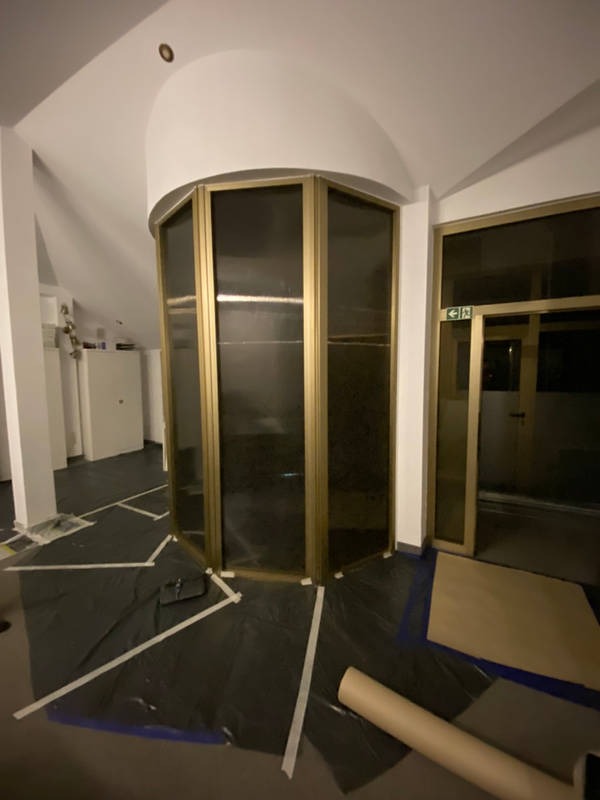 Przygotowanie do prac wykończeniowych: pomieszczenie z owalną wnęką i przeszkloną konstrukcją, podłoga zabezpieczona czarną folią i taśmą malarską, widoczny kartonowy rulon.