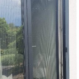 Pracownia Dekoracji Okien i Wnętrz - Złożona plisowana siatka przeciw owadom w szarej ramie, zamontowana na drzwiach balkonowych, widok z zewnątrz.