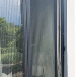 Złożona plisowana siatka przeciw owadom w szarej ramie, zamontowana na drzwiach balkonowych, widok z zewnątrz.