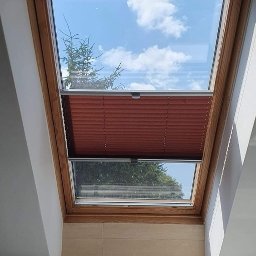 PRACOWNIA DEKORACJI OKIEN I WNĘTRZ AGNIESZKA DEREK - Brązowa plisa zamontowana w drewnianej ramie okna dachowego; widok na błękitne niebo z chmurami i zielone drzewo.