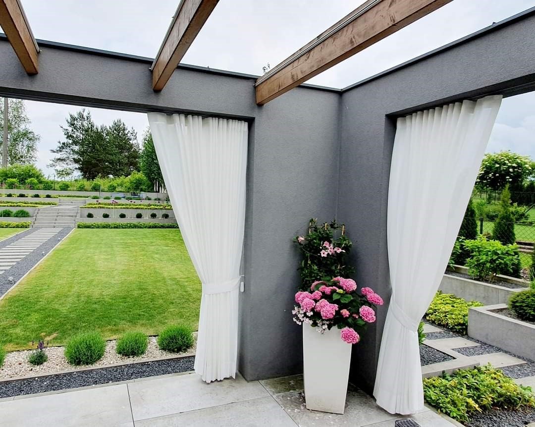 Białe, zwiewne zasłony na zewnątrz altany z widokiem na zadbany ogród z kaskadowymi rabatami i ścieżką z płyt.