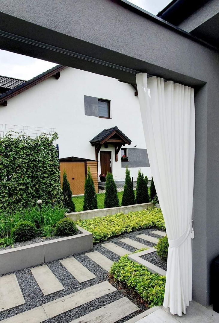 Nowoczesny ogród z minimalistycznym chodnikiem z płyt i żwiru, ozdobnymi krzewami i żywopłotem, widziany spod zadaszenia z powiewną białą zasłoną.