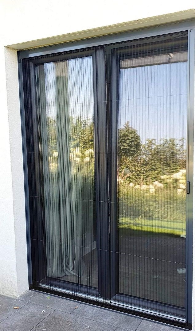 Zewnętrzna moskitiera plisowana w kolorze antracytowym zamontowana na oknie tarasowym, widok z bliska na tle zieleni ogrodu.