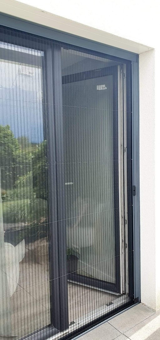 Złożona plisowana siatka przeciw owadom w szarej ramie, zamontowana na drzwiach balkonowych, widok z zewnątrz.