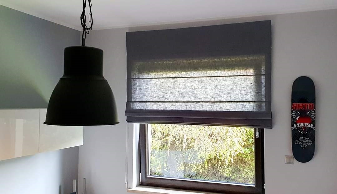 Szara roleta rzymska zamontowana w oknie, widok na zieleń za oknem, obok wisi czarna lampa i deskorolka.