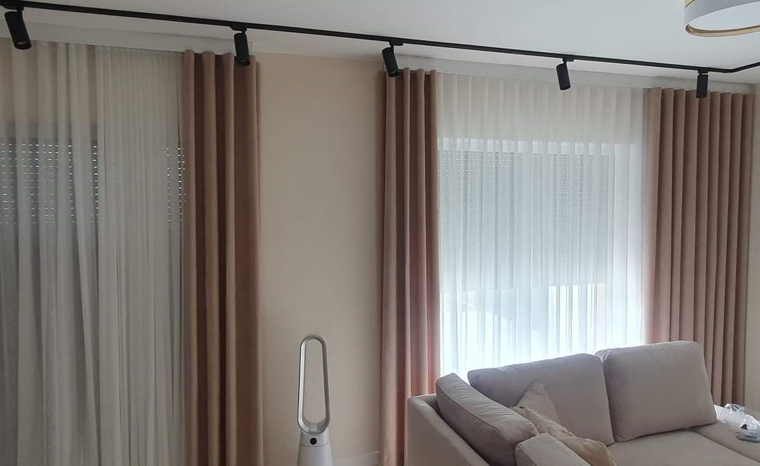 Salon z beżowymi zasłonami zaciemniającymi i białymi firanami, oświetlonymi czarnym systemem szynowym z reflektorami punktowymi.