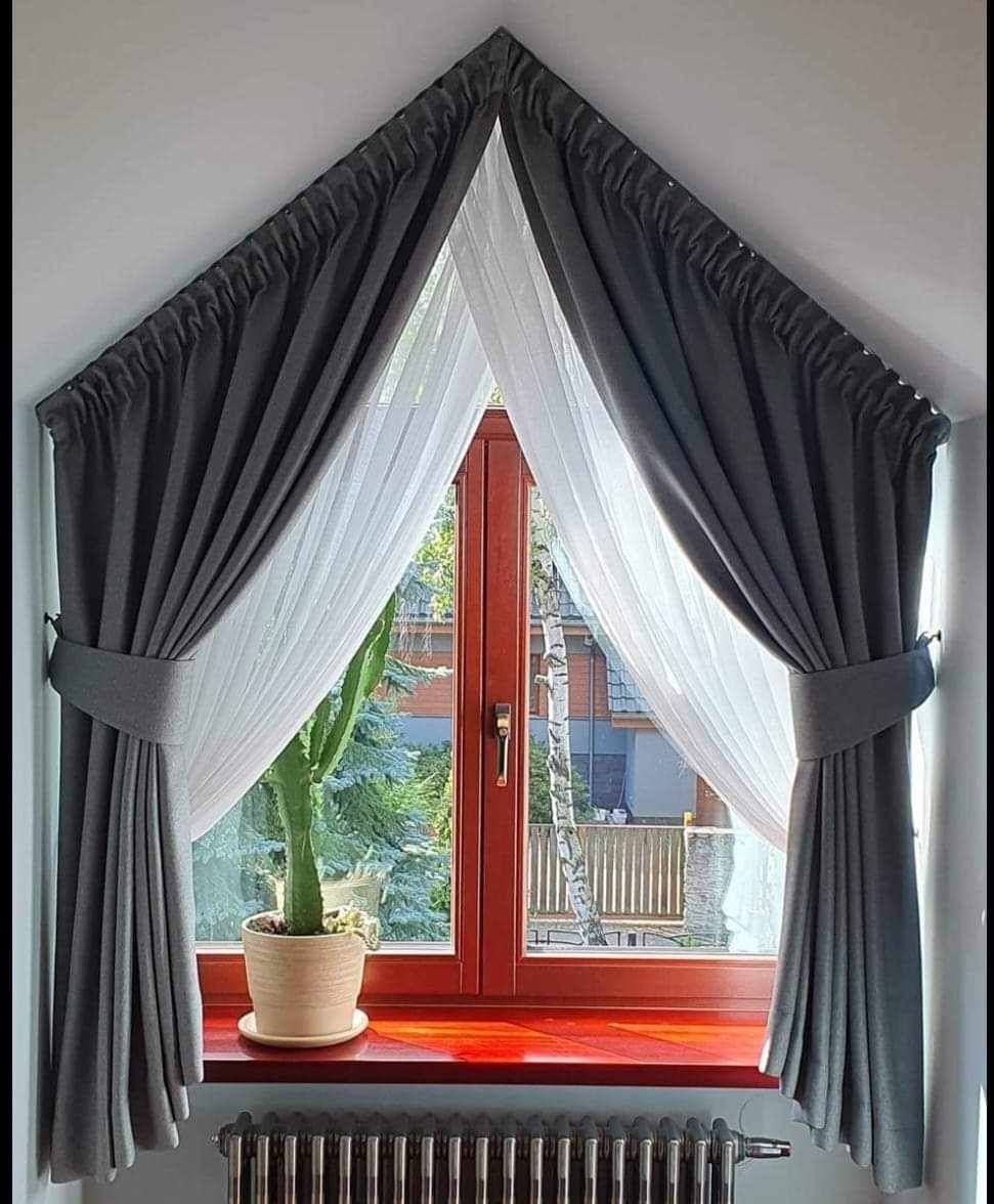 Okno trójkątne z szarymi zasłonami upiętymi pasami i białą firaną, na parapecie kaktus w doniczce, widok na zewnątrz na dom i drzewa.