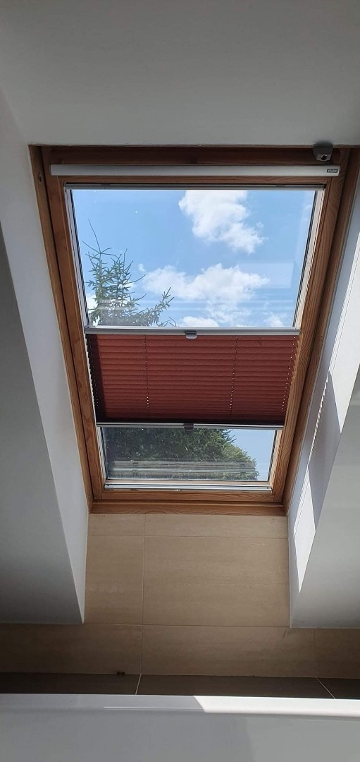 Brązowa plisa zamontowana w drewnianej ramie okna dachowego; widok na błękitne niebo z chmurami i zielone drzewo.