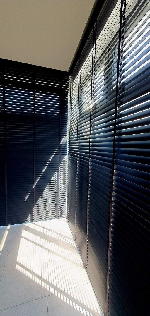 Ciemne drewniane żaluzje poziome zamontowane w rogu pokoju, rzucające równoległe cienie na jasną podłogę.