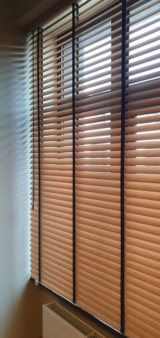 Zamknięte drewniane żaluzje poziome z ciemnymi sznurkami na oknie, widoczne z bliska, z delikatnym światłem wpadającym przez szpary.