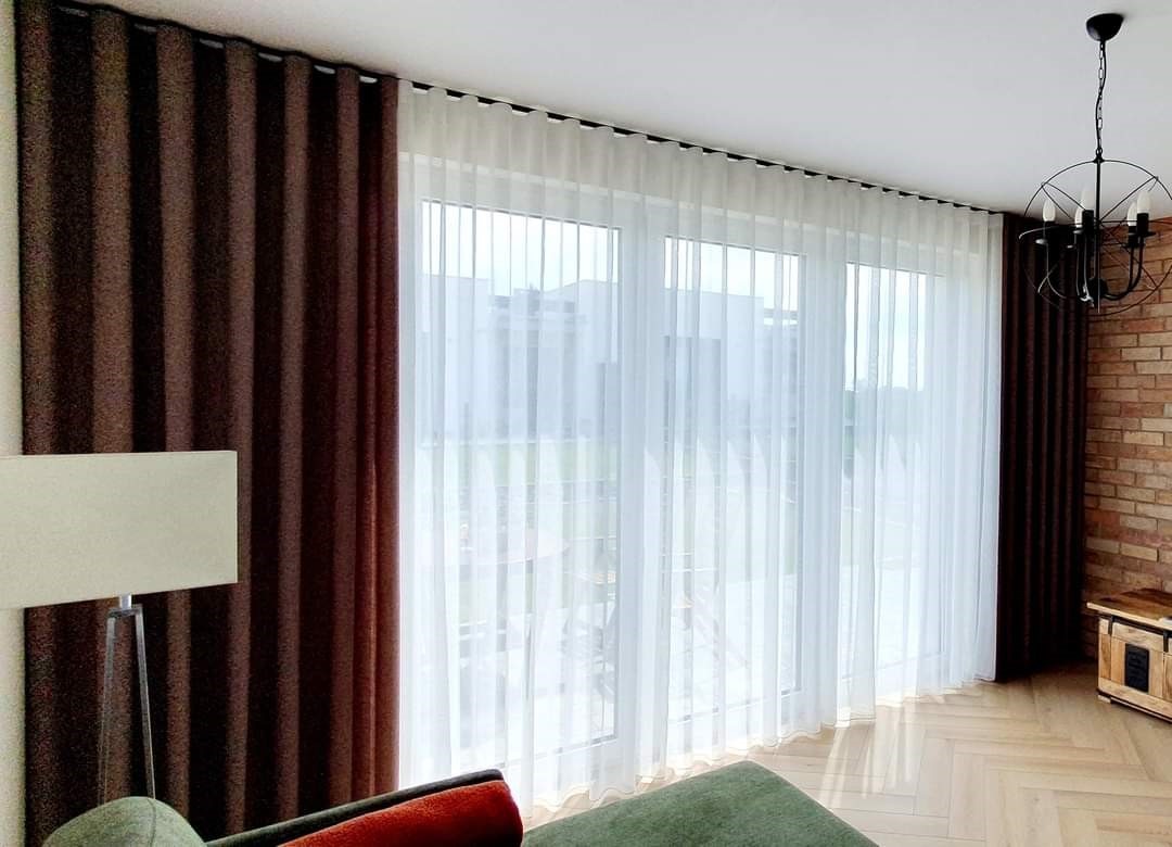 Eleganckie, brązowe zasłony zaciemniające i delikatne, białe firany zdobiące okno balkonowe w nowoczesnym salonie z ceglaną ścianą i designerską lampą.