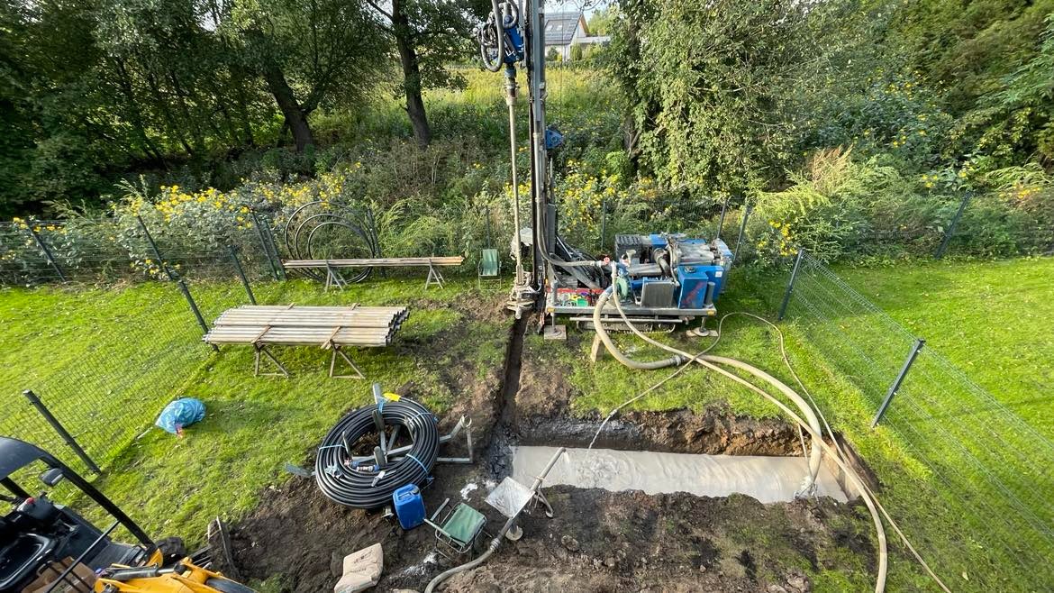 Wiertnica w trakcie wykonywania odwiertu pionowego na zielonej działce, widoczne rury, przewody, wykop i narzędzia.