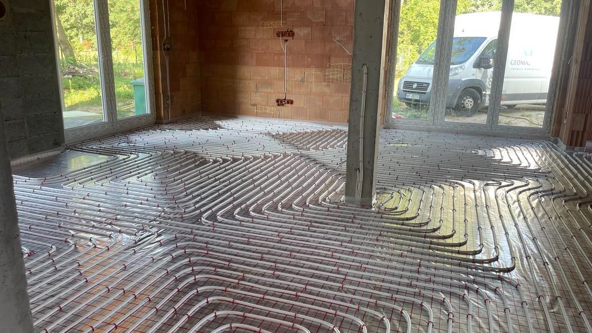 Instalacja ogrzewania podłogowego w surowym pomieszczeniu z widoczną instalacją rurową na siatce, w tle częściowo widoczny samochód dostawczy firmy Geomal.