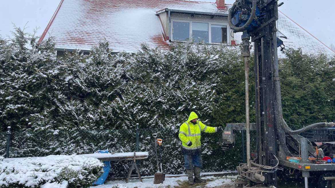 Pracownik w odblaskowej kurtce obsługuje wiertnicę do odwiertów geotermalnych na tle ośnieżonych krzewów i domu w zimowy dzień.