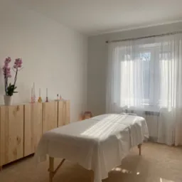 Jasne wnętrze gabinetu z drewnianą komodą, orchideą w doniczce i łóżkiem do zabiegów przykrytym białym prześcieradłem, oświetlone promieniami słonecznymi wpadającymi przez okno z firanką.