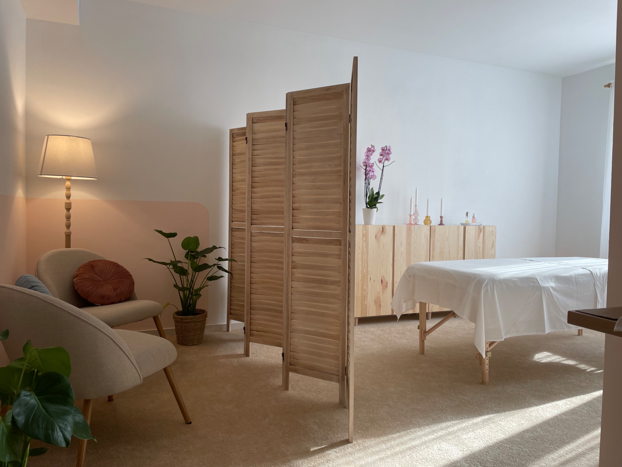 Jasne wnętrze gabinetu z drewnianym parawanem, łóżkiem do zabiegów przykrytym białym prześcieradłem, komodą z dekoracjami (świeczki, storczyk) oraz dwoma fotelami z okrągłą poduszką i roślinami...