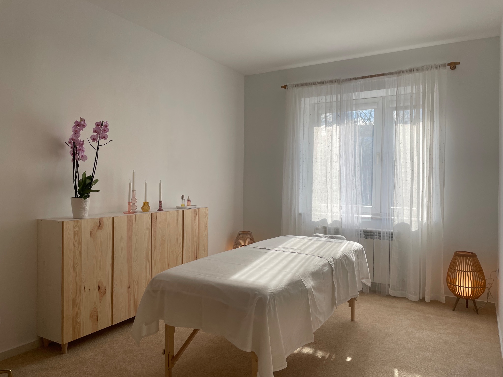 Jasne wnętrze gabinetu z drewnianą komodą, orchideą w doniczce i łóżkiem do zabiegów przykrytym białym prześcieradłem, oświetlone promieniami słonecznymi wpadającymi przez okno z firanką.