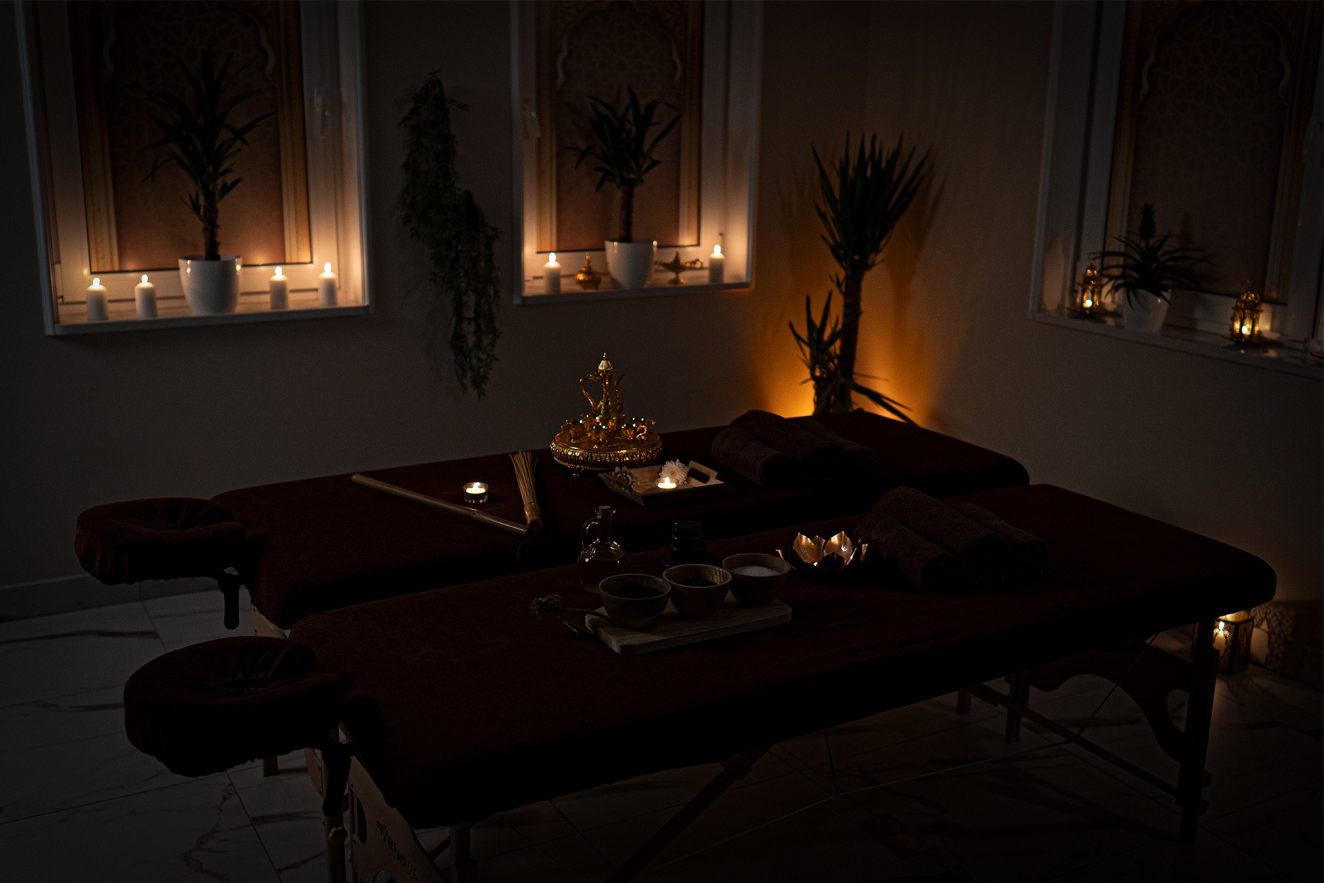 Dwa ciemne łóżka do masażu w zaciemnionym pokoju, dekorowane świecami, roślinami doniczkowymi i orientalnymi naczyniami.